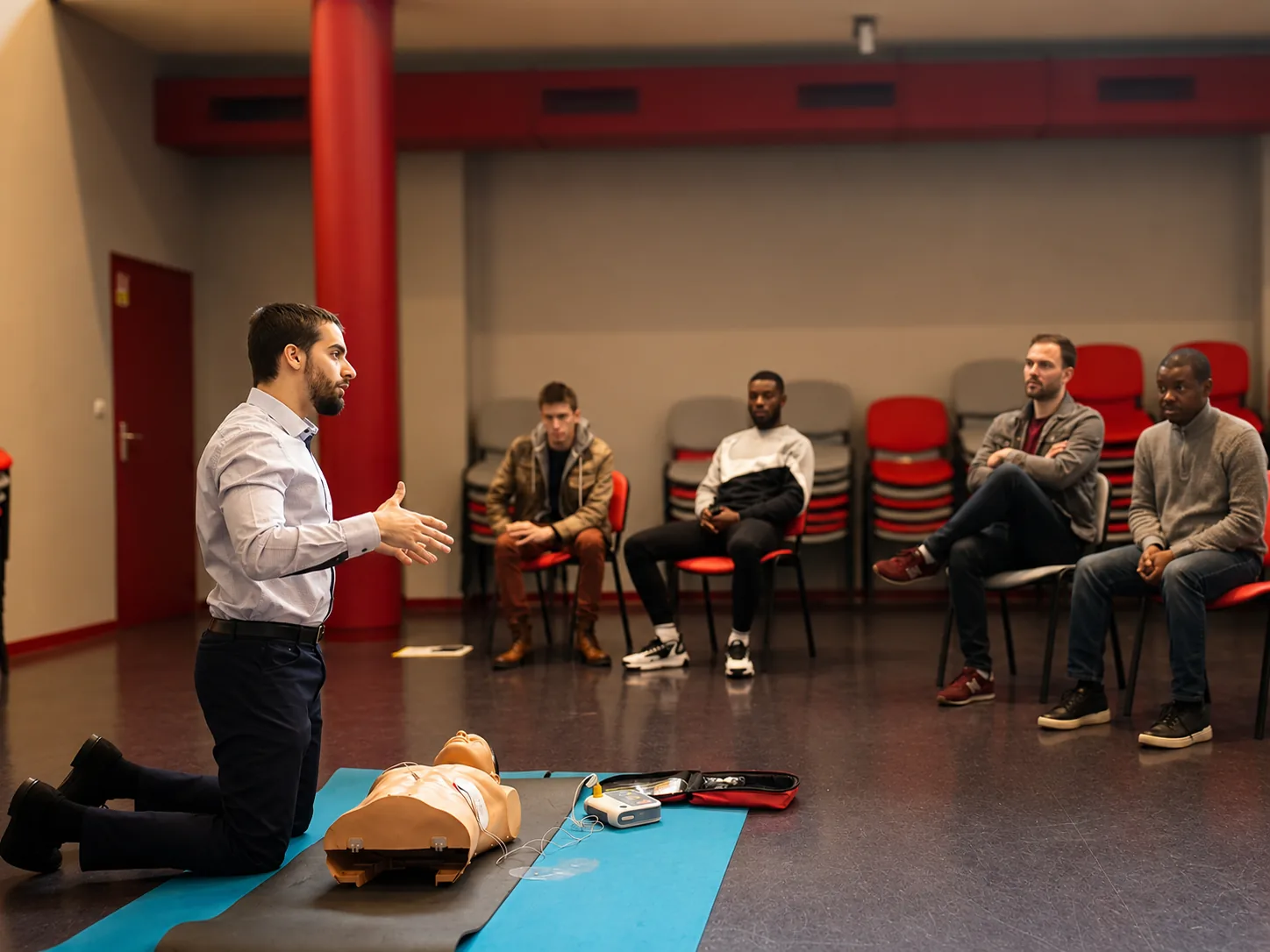 Formateur Red Life Academy animant une session de secourisme devant un groupe, mannequin de RCP et défibrillateur au sol